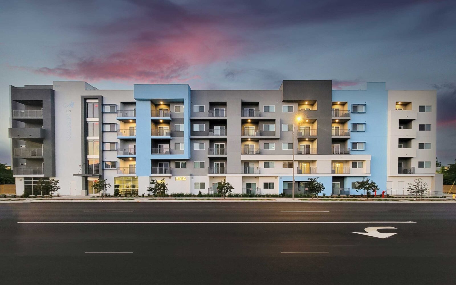 A modern apartment building with a gray and blue façade stands across a street under a twilight sky with pink clouds. Some balcony lights are on, and young trees are planted near the sidewalk.